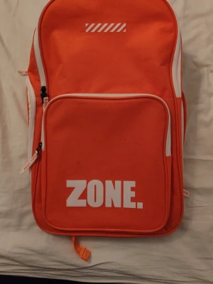 Orange ryggsäck från Zone - Snygg orange ryggsäck från Zone med vit logga och detaljer. Ryggsäcken har ett stort huvudfack, en stor framficka med dragkedja och vadderade axelband med Zone-tryck. Tillverkad i slitstark textil och har vita dragkedjor som kontrast. Perfekt för innebandyspelare och även för vardagslivet. Hör av er vid frågor. Pris kan diskuteras vid snabb affär!😀