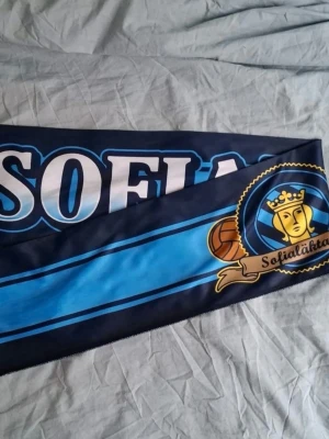Sofialäktaren duk pris diskuteras - Snygg supporterhalsduk i blått och svart med texten 'Sofialäktaren' och klubbmärke med guldkrona och fotboll. Perfekt accessoar för dig som vill visa ditt stöd på matchen.