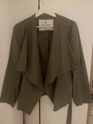 Olivgrön trenchcoat från Chiara Forthi - Snygg olivgrön trenchcoat från Chiara Forthi Milano med draperad krage och knytskärp i midjan. Jackan har långa ärmar och en stilren, modern design som passar perfekt till olika outfits. Materialet är mjukt och lätt, vilket gör den bekväm att bära.