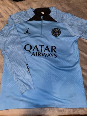 PSG träningströja blå långärmad - Snygg blå långärmad träningströja från Jordan x PSG med svart detalj vid halsen och halv dragkedja. Tröjan har PSG-logga och Jordan-logga på bröstet samt Qatar Airways-tryck framtill. Tillverkad i lätt och ventilerande material, perfekt för fotbollsträning.