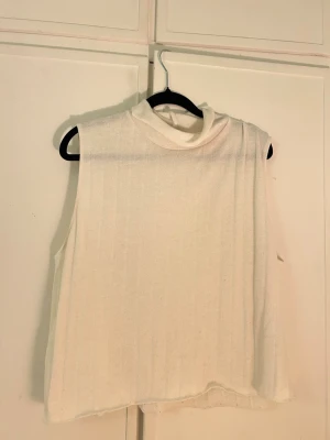 Vit ribbad ärmlös polotopp - Säljer en vit ärmlös polotopp med ribbad struktur. Toppen har hög krage och är gjord i ett mjukt material som känns skönt mot huden. Perfekt att styla med jeans eller kjol för en clean look.