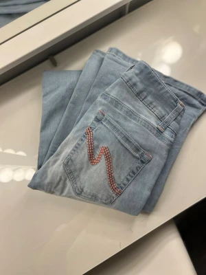 Ljusblå jeans från Nelly med brodyr - Säljer ett par ljusblå jeans från Nelly med snygg röd brodyr på bakfickan. Jeansen har klassisk femficksmodell, dubbla knappar i midjan och raka ben. Perfekta för dig som gillar en avslappnad men trendig stil.