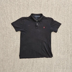 Svart pikétröja från Polo Ralph Lauren - Svart pikétröja från Polo Ralph Lauren med klassisk krage, två vita knappar och röd broderad logga på bröstet. Tillverkad i mjuk bomull och har korta ärmar för en avslappnad men stilren look. Har en väldigt fadad vita fläck vid kragen som lätt går bort i tvätt