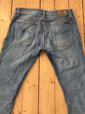 Ljusblå jeans slim från Nudie Jeans - Säljer ett par ljusblå jeans från Nudie Jeans med klassisk femficksdesign och raka ben. Jeansen har snygga slitningar och kontrastsömmar samt Nudie-loggan på bakfickan. Perfekta för en avslappnad och trendig look.