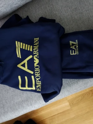 EA7 / Emporio Armani Tracksuit – Hoodie + Mjukisbyxor (Mörkblå) - Säljer ett snyggt och bekvämt set från Emporio Armani / EA7. Setet består av en mörkblå hoodie och matchande mjukisbyxor med guldfärgad EA7-logga. Väldigt stilrent och sportigt, passar både till vardags och för en mer avslappnad stil. Hoodien har huva och en stor ficka framtill, medan byxorna har resår i midjan och vid bensluten för en bra passform. Materialet är mjukt och skönt att bära. Storlek S i vuxen storlek så passar barn som har upp till storlek L. Det är i nyskick och säljes som set