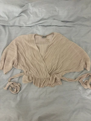 Beige omlottopp med knytband - Trendig beige omlottopp med knytband i midjan och korta ärmar. Toppen har ett ribbat tyg och croppad passform som ger en modern look. Perfekt att styla med höga jeans eller kjol. Mer champagne i verkligheten 