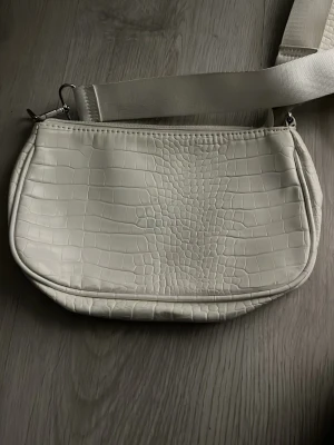 Vit axelväska med krokoprägling - Snygg vit axelväska i fejkläder med krokodilpräglad yta och bred axelrem. Väskan har en rund liten matchande coin purse som sitter fast i remmen. Perfekt för dig som gillar trendiga accessoarer med lite extra detalj.