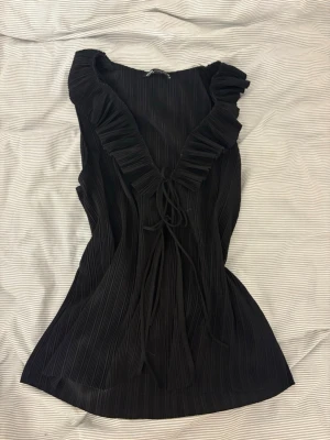 Svart plisserad topp från Zara - Säljer en svart plisserad topp från Zara med djup v-ringning och volangkant. Toppen har knyt framtill och är ärmlös, vilket ger en trendig och luftig känsla. Perfekt för dig som gillar detaljer och en chic look.