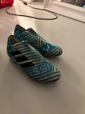 Adidas Messi blå fotbollsskor storlek 39 1/3 - Säljer ett par blåa Adidas Messi fotbollsskor med vita och svarta detaljer. Skorna har ett mönstrat textilöverdrag och snörlös design för tight passform. Messi-logga och Adidas-ränder på sidorna.