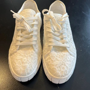 Sommmriga vita sneakers - Snygga vita sneakers med blommig spets över hela skon och vita snören. Skorna har en platt sula och rund tå, vilket ger en fräsch och trendig look. Perfekta för dig som vill ha något unikt och stilrent till din outfit.Är använda 1 gång.De ser mer beige ut på bilden pga ljuset, men är vita!!Nypris: 900 kr