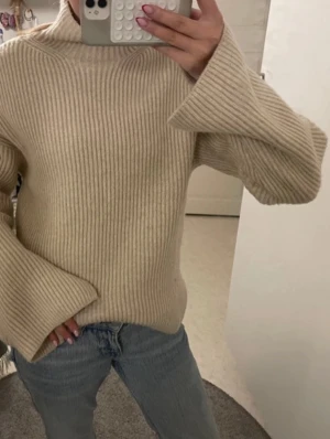 Stickad tröja - Säljer en beige ribbstickad tröja med höghals och extra vida ärmar. Tröjan har en oversized passform och är gjord i ett mjukt, tjockt material som känns riktigt cozy. Perfekt för lager på lager och chill dagar.
