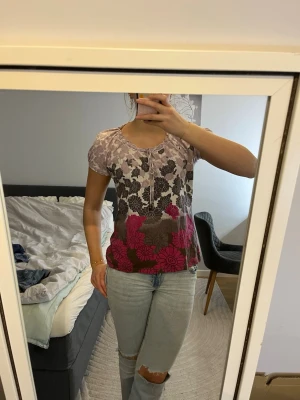 Blommig topp med snörning - Säljer en blommig topp med snörning framtill. Toppen har korta ärmar och ett mönster i ljus rosa,grå, vit och lila färg med stora blommor. Materialet känns lätt och bekvämt, perfekt för dig som gillar färg och mönster.