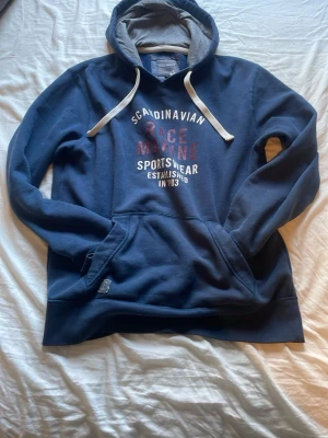 Blå hoodie från Race Marine - Mörkblå hoodie från Race Marine med vit och röd text på bröstet. Stor känguruficka framtill, vita dragsnören och grå insida i huvan. Tillverkad i mjuk bomullsblandning, perfekt för en avslappnad stil.