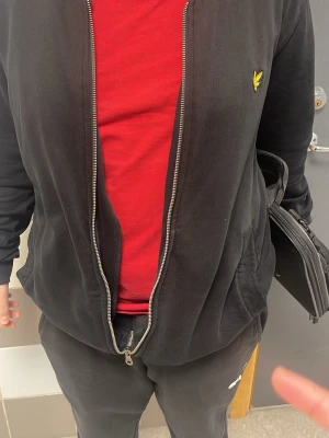 Svart hoodie från Lyle & Scott - Svart hoodie med dragkedja från Lyle & Scott, prydd med den klassiska gula örnloggan på bröstet. Hoodien har ribbade muddar och fickor på sidorna. Perfekt för dig som vill ha en stilren och bekväm look.