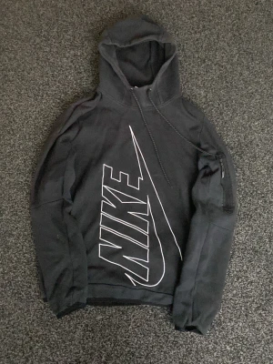 Svart hoodie från Nike med stor logga - Säljer en svart hoodie från Nike med en stor vit logga och swoosh på framsidan. Hoodien har huva med dragsko och en ficka på ärmen med dragkedja. Tillverkad i mjukt bomullsmaterial, perfekt för en avslappnad streetwear-look.