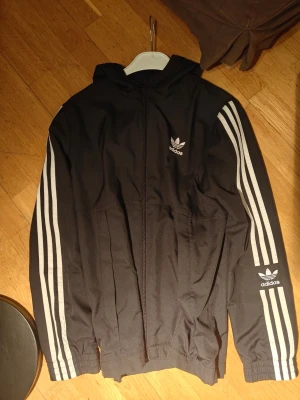 Svart Adidas vindjacka med huva - Snygg svart vindjacka från Adidas med klassiska vita ränder längs ärmarna och Adidas Trefoil-logga på bröstet. Jackan har dragkedja framtill, två sidofickor och huva. Tillverkad i lätt och tåligt material, perfekt för en sportig och avslappnad stil.