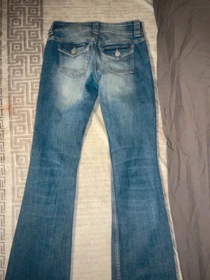 Blå bootcut jeans med ficklock - Säljer ett par blå bootcut jeans med snygg slitning och ficklock bak med knappdetaljer. Jeansen har breda benslut och klassisk femficksdesign. Perfekta för dig som gillar en retro vibe och vill ha ett par jeans som sticker ut lite extra.