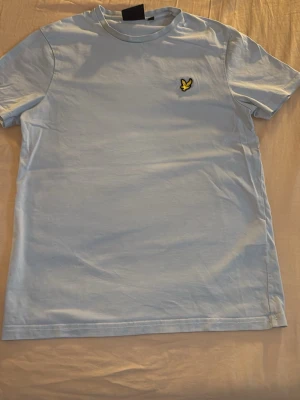 Ljusblå t-shirt från Lyle & Scott - Snygg ljusblå t-shirt från Lyle & Scott med klassisk gul logga på bröstet. T-shirten har rund halsringning och korta ärmar. Tillverkad i mjuk bomull för skön känsla och enkel stil.