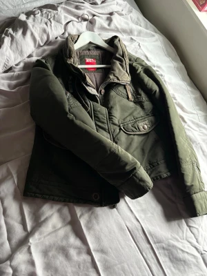 Grön jacka från Miss Sixty - Snygg grön jacka från Miss Sixty med hög krage, dragkedja och knappar framtill. Jackan har flera fickor och coola detaljer som snörning vid kragen. Perfekt för dig som vill ha en stilren och funktionell look.