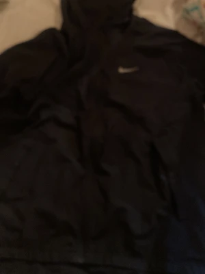 Svart Nike vindjacka med huva - Snygg svart vindjacka från Nike med huva och dragkedja framtill. Jackan har en diskret Nike-logga på bröstet och är tillverkad i lätt, vindtätt material. Perfekt för löpning eller promenader när det blåser.