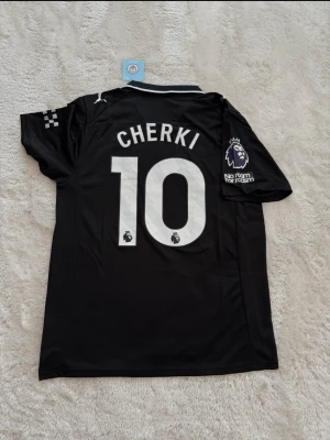 Svart Manchester City tröja Puma - Snygg svart Manchester City matchtröja från Puma med CHERKI och nummer 10 på ryggen. Tröjan har Premier League-logga, Etihad Airways-tryck på bröstet och detaljer i vitt. Perfekt för fotbollsfans som vill sticka ut på planen eller läktaren.