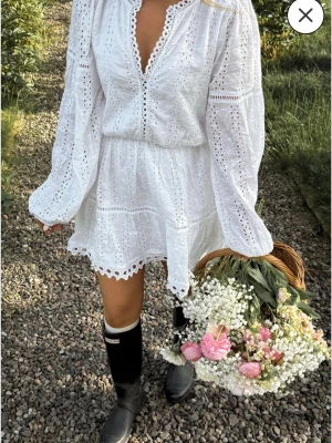 Vit broderad kort klänning med spets - Superfin vit kort klänning med broderade detaljer och spetskant. Klänningen har v-ringning, långa ärmar och markerad midja. Perfekt för sommarens alla tillfällen och ger en romantisk vibe.