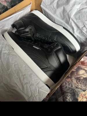 Svarta Puma sneakers med vit sula - Stilrena svarta sneakers från Puma med klassisk vit sula. Skorna har en låg profil, snygga sömdetaljer och är tillverkade i slätt lädermaterial. Puma-loggan syns på sidan och de har svarta skosnören för en clean look. 