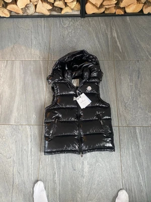 Svart Moncler väst  - . Stilren och lyxig svart dunväst från Moncler med högblank finish. Västen har en vadderad design som ger både värme och en modern look. Utrustad med huva, dragkedja framtill och två sidofickor med dragkedja. Den ikoniska Moncler-loggan sitter på bröstet och ger en exklusiv touch. Perfekt för kyliga dagar när du vill kombinera funktion och stil.  