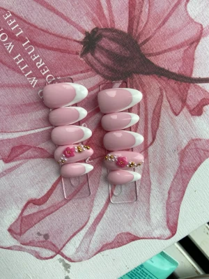 Rosa press-on naglar med blommor - Snygga press-on naglar i ljusrosa med vit fransk tipp. Några naglar har dekorativa rosa blommor och små guldfärgade pärlor. Ovala och glansiga, perfekta för dig som vill ha en söt och trendig look.