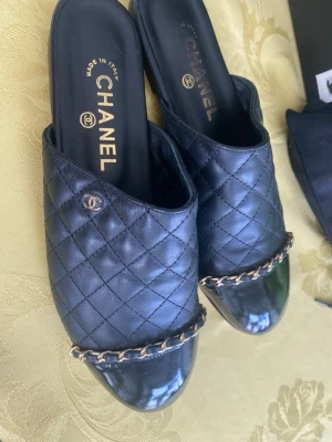 Svarta quiltade tofflor från Chanel - Säljer ett par svarta tofflor från Chanel i quiltat läder med guldfärgad kedjedetalj framtill och diskret logga på sidan. Tillverkade i Italien och har en klassisk, lyxig look som passar perfekt för dig som gillar exklusiva accessoarer. Märkta strl 38. Endast provade en gån inomhus. Plast sitter kvar på skinnsulan.
