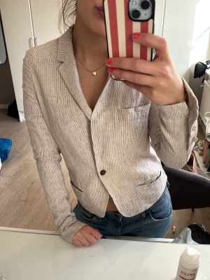 Beige smalrandig kavaj - Snygg beige kavaj med smala vita ränder och klassisk krage. Två knappar framtill och fickdetaljer. Figurnära passform som ger en stilren look. Perfekt att matcha med jeans för en avslappnad men uppklädd stil.