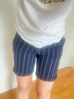Randiga blå shorts med hög midja - Snygga blå shorts med vita vertikala ränder och hög midja. Shortsen har bakficka med knapp och är perfekta för en trendig sommarlook. Sköna och lätta att matcha med olika toppar.