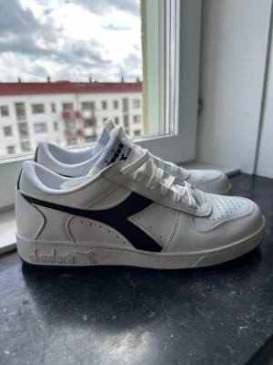 Diadora Magic Basket Icona sneakers - Snygga vita sneakers från Diadora med svart logga på sidan. Skorna har klassisk låg profil, perforerad tå och vita skosnören. Tillverkade i slätt läder med bekväm vadderad kant och sula i gummi. Perfekta för en clean och sportig stil.