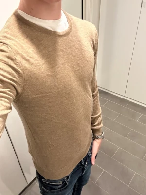 Beige stickad tröja i ullmix - Snygg beige stickad tröja med rund halsringning och långa ärmar. Tröjan har en enkel och stilren design som passar till mycket. Materialet känns mjukt och lätt, troligen en ull- eller bomullsblandning. Perfekt att bära över en t-shirt för en avslappnad look.