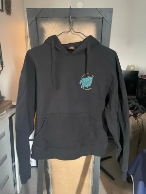 Svart Santa Cruz hoodie med tryck - Säljer en svart hoodie från Santa Cruz med turkost och gult logotryck på bröstet och ryggen. Klassisk känguruficka framtill och justerbar huva med snören. Perfekt för dig som gillar streetwear och vill ha en snygg och bekväm hoodie.