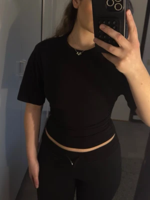 Svart croppad topp med snörning - Snygg svart croppad topp med korta ärmar och cool snörningsdetalj i sidan. Toppen har en tight passform och är tillverkad i mjukt bomullsmaterial. Perfekt till jeans eller kjol för en trendig look.