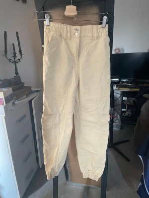 Beige cargobyxor från Bershka - Snygga beige cargobyxor med resår i midjan och vid bensluten. Byxorna har flera praktiska fickor på sidorna och bak, samt dubbla knappar framtill, perfekta för en avslappnad streetwear-look.