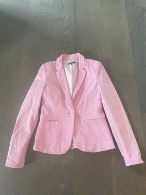 Ljusrosa figursydd kavaj - Snygg ljusrosa kavaj med figurnära passform och klassisk krage. Kavajen har en knapp framtill och två fickor. Perfekt för att lyfta vilken outfit som helst med en poppig färg. Materialet ser ut att vara bomullsblandning.