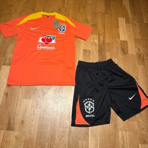 Nike Brasil summer set orange & svart - Säljer ett Brasil träningsset från Nike med orange t-shirt och svarta shorts. Tröjan har gula detaljer, tryckt Brasil-märke och Guaraná sponsorlogga. Shortsen har Brasil-logga och orange detalj. Perfekt för fotbollsträning eller gym. Materialet är lätt och andas.