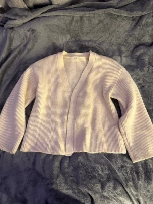 Vit ribbstickad kofta med v-ringning - Mysig vit ribbstickad kofta med v-ringning och raka ärmar. Koftan har en avslappnad passform och är tillverkad i ett mjukt material som känns skönt mot huden. Perfekt att slänga över en t-shirt eller topp för en enkel och stilren look.