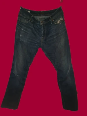 Jack&Jones  - Säljer ett par mörkblå jeans med rak passform och råa, avklippta benslut. Jeansen har klassiska fem fickor och slitna detaljer framtill. Perfekta för en avslappnad och trendig look.