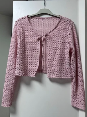 Rosa virkad kofta med rosett - Söt ljusrosa virkad kofta med lång ärm och knytband framtill. Koftan är kortare i modellen och har ett luftigt, genombrutet mönster som ger en romantisk vibe. Perfekt att slänga över ett linne eller klänning för en extra touch.