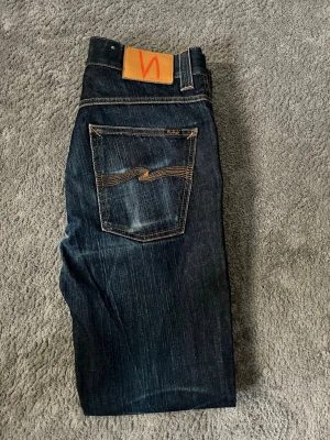 Nudie Jeans mörkblåa dry Japan  - Säljer ett par mörkblå jeans från Nudie Jeans med klassisk femficksmodell och kontrastsömmar. Modellen är dry Japan low slim Jim med bootcut. Jeansen har en snygg tvätt och den ikoniska Nudie-bågen på bakfickorna. Skick 7/10 pågrund av ett hål som syns på bilderna nypris - 2500 säljs - 1149kr! Vid minsta fundering bara att höra av sig!☑️