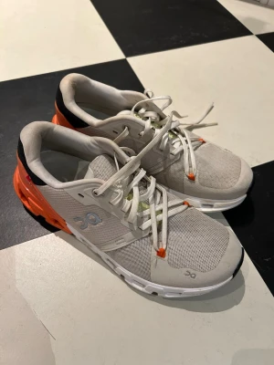 CloudFlyer från On - Snygga sneakers från On i vitt meshmaterial med orange och svarta detaljer. Skorna har vita snören, ventilerande ovandel och en chunky sula med On-logga på sidan. Perfekta för dig som gillar sportig stil och vill ha bekväma skor till träning, löpning eller vardag.