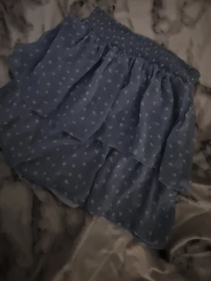 Blå volangkjol med små vita hjärtan - Supersöt blå kortkjol med volanger och små vita hjärtan. Kjolen har resår i midjan och är gjord i ett lätt, luftigt material som passar perfekt till varmare dagar.