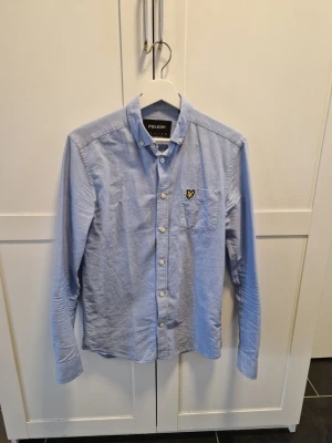 Ljusblå skjorta från Lyle & Scott - Klassisk ljusblå skjorta från Lyle & Scott med knappslå och vit knappstängning. Skjortan har lång ärm och en bröstficka med den ikoniska gula loggan. Perfekt till jeans eller chinos för en clean och stilren look.
