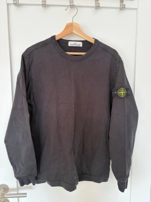 Svart sweatshirt från Stone Island - Svart långärmad sweatshirt från Stone Island med klassisk rund halsringning och ikonisk logotyp-patch på vänster ärm. Tröjan har en avslappnad passform och är tillverkad i mjuk bomull, perfekt för en streetwear-look.