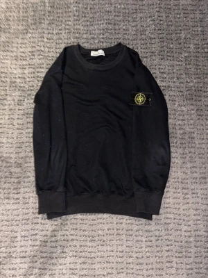 Svart sweatshirt från Stone Island - Svart sweatshirt från Stone Island med den ikoniska kompass-loggan på vänster ärm. Tröjan har rund halsringning, ribbade muddar vid ärmslut och nederkant samt en clean och stilren look. Perfekt för dig som gillar streetwear och vill ha något klassiskt.