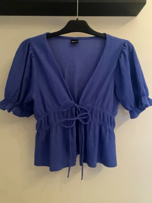 Blå topp med puffärm och knyt - Supersöt blå omlottopp med puffiga korta ärmar och knyt i midjan. Toppen har v-ringning och lätt rynkad midja som ger en feminin siluett. Perfekt att styla med jeans eller kjol.  Superfint skick och roligt med en topp med färg💙