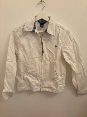 Vit jacka från Polo Ralph Lauren - Klassisk vit jacka från Polo Ralph Lauren med dragkedja framtill och två fickor. Jackan har en krage med rutigt foder i blått och rött samt den ikoniska loggan broderad på bröstet. Perfekt för dig som gillar stilrena och tidlösa plagg.